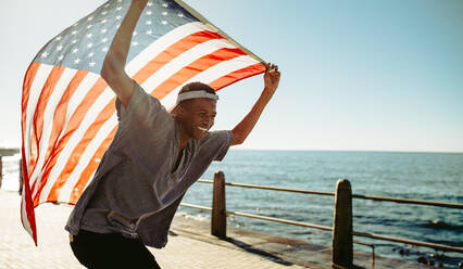 Lächelnder afrikanischer Mann an der Strandpromenade mit amerikanischer Flagge. Fröhlicher junger Mann an der Promenade mit amerikanischer Flagge. - JLPSF18053