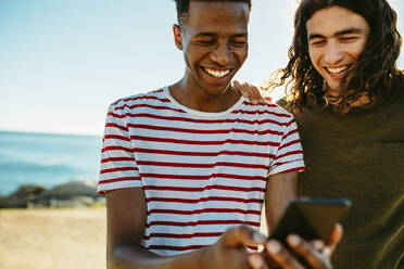 Zwei junge Freunde im Freien, die auf ihr Handy schauen und lächeln. Junge Männer benutzen ein Smartphone am Strand. - JLPSF18054