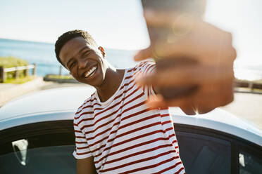 Lächelnder junger afrikanischer Mann, der ein Selfie macht, während er neben einem Auto im Freien steht. Afroamerikanischer Mann, der ein Selbstporträt im Freien an einem Sommertag macht. - JLPSF18055