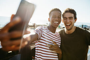 Zwei lächelnde junge Männer machen ein Selfie, während sie an einem Auto im Freien stehen. Freunde machen ein Selbstporträt im Freien an einem Sommertag. - JLPSF18056