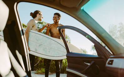 Shot from inside a car of two men standing outside with surfboards. Male friends on a surfing holiday standing by a car. - JLPSF18063