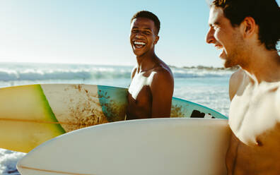 Fröhlicher junger Mann mit einem Freund, der mit Surfbrettern am Strand spazieren geht. Fröhliche junge Freunde im Urlaub am Meer. - JLPSF18065
