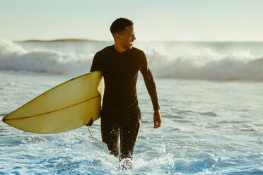 Lächelnder junger Mann mit Surfbrett am Strand. Afrikanischer Mann, der nach dem Wassersurfen aus dem Meer kommt. - JLPSF18070