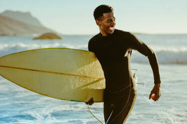 Lächelnder afrikanischer Mann, der nach dem Wassersurfen aus dem Meer läuft. Glücklicher junger Mann mit Surfbrett am Strand, der seinen Urlaub genießt. - JLPSF18071