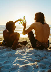 Rückansicht von zwei jungen Freunden, die bei Sonnenuntergang am Strand sitzen und mit Bier anstoßen. Junge Leute, die am Strand mit Bier feiern. - JLPSF18074