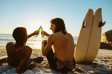 Rückansicht von zwei jungen Surfern, die am Strand auf ein Bier anstoßen. Junge Männer feiern am Strand mit Surfbrett an der Seite. - JLPSF18075