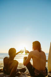 Rückansicht von zwei jungen Männern, die sich mit Bier am Strand entspannen. Junge Männer stoßen bei Sonnenuntergang mit Bier am Strand an. - JLPSF18076