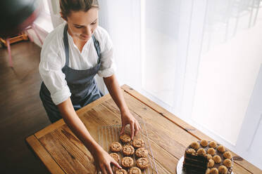 Draufsicht auf eine Konditorin, die in der Küche Kekse zubereitet. Eine Bäckerin arrangiert Kekse auf einem Grill mit einem Kuchen auf dem Tisch. - JLPSF18084