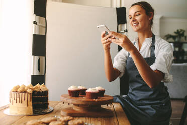 Glückliche Frau Koch ein Bild von Gebäck auf einem Holzbrett mit Smartphone, während in der Küche stehend. Weibliche Bäckerin, die Bilder des Desserts für ihre Lebensmittel-Blog. - JLPSF18088