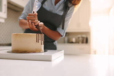 Frau in Schürze dekoriert den Kuchen mit flüssiger Schokolade. Konditorin in der Küche dekoriert Kuchen mit Schokoladenglasur. - JLPSF18101