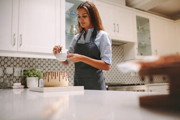 Frau mischt flüssige Schokolade in einer Schüssel, um den Kuchen zu dekorieren. Frau in Schürze bereitet Schokoladenkuchen zu Hause vor. - JLPSF18108