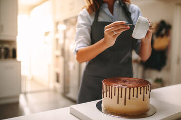 Konditorin in der Küche, die einen Schokoladenkuchen dekoriert. Frau in Schürze gießt flüssige Schokolade über einen Kuchen auf dem Küchentisch. - JLPSF18112