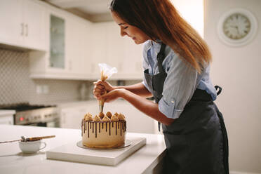 Köchin dekoriert Kuchen mit Schlagsahne mit Partytüte. Frau in Schürze bereitet einen leckeren Kuchen zu Hause vor. - JLPSF18117