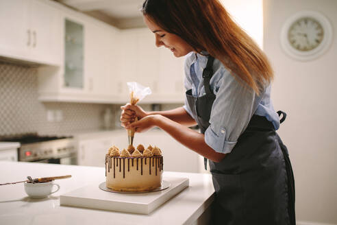 Female chef decorating cake with whipped cream using party bag. Woman in apron preparing a delicious cake at home. - JLPSF18117