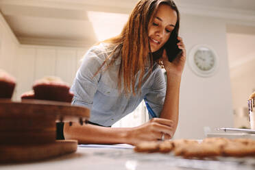 Konditorin, die eine Bestellung aufschreibt und mit einem Smartphone spricht. Konditorin, die in der Küche Bestellungen per Telefon entgegennimmt. - JLPSF18125