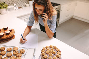 Draufsicht auf eine Konditorin, die in der Küche Bestellungen per Telefon entgegennimmt. Eine Köchin schreibt eine Bestellung, während sie mit dem Handy telefoniert. - JLPSF18126