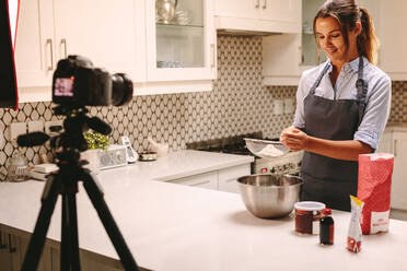 Video camera filming young female blogger at the kitchen. Woman pastry chef sifting flour in a bowl to make dough with a digital camera recording the session for a food video blog. - JLPSF18138