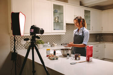 Young woman wearing apron filming herself preparing cake in the kitchen. Pastry chef recording content for the food and baking vlog using a camera mounted on tripod. - JLPSF18143