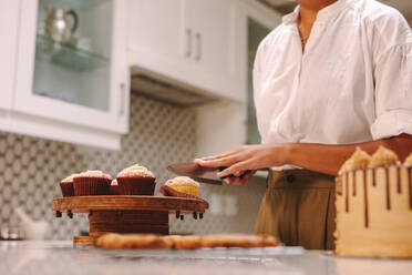 Weibliche Köchin schneidet einen Cupcake auf Holzbrett mit Messer in der Küche. Frau Konditorin arbeitet in der Küche. - JLPSF18147