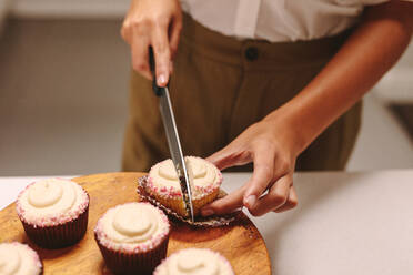 Close up of pastry chef cutting a homemade cupcake on wooden board with knife. Woman confectioner preparing muffins in the kitchen. - JLPSF18148