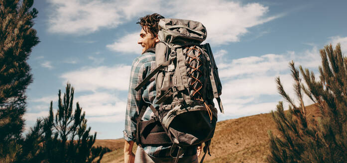 Rear view shot of happy young man with backpack going on a camping. Caucasian male hiker on mountain looking away and smiling. - JLPSF18167