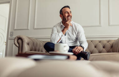 Porträt eines lächelnden Geschäftsmannes, der auf dem Sofa in einem Hotelzimmer sitzt und wegschaut. Älterer Geschäftsmann, der auf einer Geschäftsreise in einem luxuriösen Hotelzimmer wohnt. - JLPSF18177