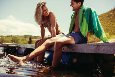 Lächelndes junges Paar sitzt auf dem Steg und lässt die Beine im Wasser baumeln. Junger Mann und Frau entspannen sich auf einem Steg am See, beide in Badekleidung. - JLPSF18188