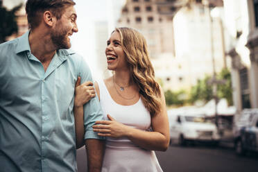 Fröhliches Paar, das lachend auf der Straße spazieren geht und sich gegenseitig anschaut. Frau hält ihren Partner, während sie auf der Straße spazieren geht. - JLPSF18199