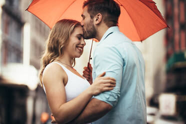 Mann hält seine Freundin und küsst sie auf die Stirn, während er auf der Straße steht. Glückliches Paar, das auf der Straße steht und sich gegenseitig umarmt und einen Regenschirm hält. - JLPSF18200