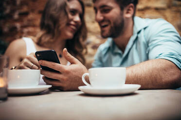 Lächelndes Paar in einem Café, das Zeit miteinander verbringt und sich unterhält. Mann sitzt mit seiner Freundin in einem Restaurant und schaut auf sein Mobiltelefon. - JLPSF18204