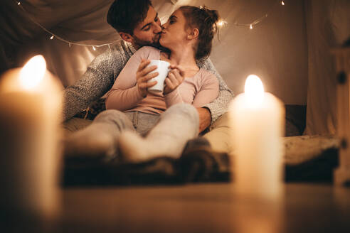 Frau, die mit ihrem Mann auf dem Bett sitzt, eine Kaffeetasse hält und ihn küsst. Verliebtes Paar, das sich küsst, während es auf dem Bett in einem mit Kerzen und kleinen Glühbirnen beleuchteten Raum sitzt. - JLPSF18206