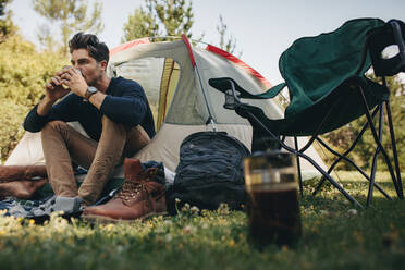 Junger Mann sitzt in seinem Zelt auf dem Campingplatz und trinkt Kaffee. Männlicher Camper entspannt sich in einem Zelt und trinkt eine Tasse Kaffee. - JLPSF18230