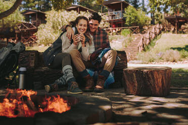 Happy young man and woman sitting by campfire with coffee. Loving couple resting by a bonfire at campsite. - JLPSF18240