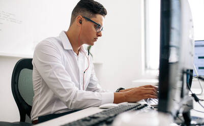 Softwareentwickler sitzt an seinem Bürotisch und arbeitet mit Kopfhörern am Laptop. Mann mit Brille arbeitet am Laptop im Büro. - JLPSF18246