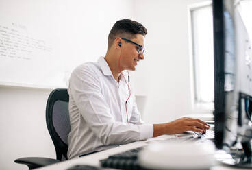 Lächelnder Softwareentwickler, der an seinem Schreibtisch sitzt und mit Kopfhörern am Laptop arbeitet. Mann mit Brille, der im Büro am Laptop arbeitet. - JLPSF18247