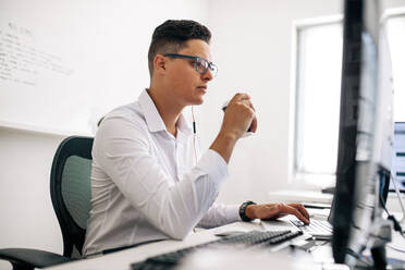 Lächelnder Softwareentwickler, der an seinem Schreibtisch sitzt und mit Kopfhörern am Laptop arbeitet. Mann mit Brille, der im Büro am Laptop arbeitet und Kaffee trinkt. - JLPSF18248