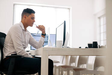 Software developer sitting at his office desk looking at laptop computer and thinking with spectacles in mouth. Man holding spectacles working on computer in office with a coffee cup on the table. - JLPSF18250