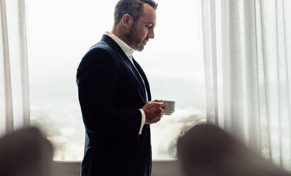 Geschäftsmann mit einer Tasse Kaffee im Hotelzimmer stehend. Reifer Mann im Anzug trinkt Kaffee im Hotelzimmer am Morgen. - JLPSF18263