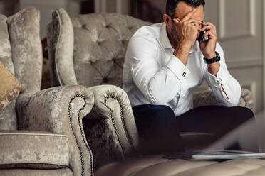 Porträt eines gestressten reifen Geschäftsmannes, der in einem Hotelzimmer sitzt und telefoniert. Geschäftsmann, der auf einer Couch sitzt und mit der Hand auf dem Kopf telefoniert. - JLPSF18268