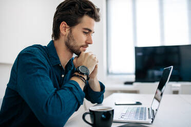 Software developer sitting in front of computer and working in office. Man looking at laptop sitting with his chin resting on hands with a coffee cup on the table. - JLPSF18269