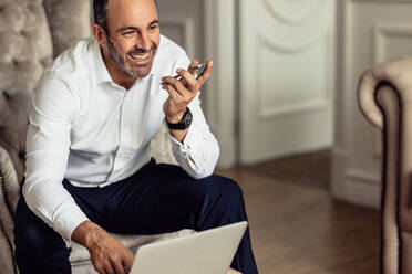 Businessman talking on phone and working on laptop while sitting in hotel room. Smiling businessman working from hotel room on business trip. - JLPSF18273