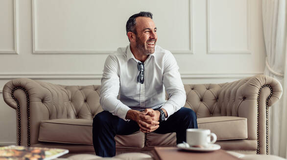 Porträt eines fröhlichen Geschäftsmannes, der auf dem Sofa in einem Hotelzimmer sitzt und lachend wegschaut. CEO, der in einem luxuriösen Hotelzimmer auf Geschäftsreise ist. - JLPSF18275