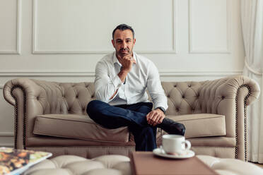 Portrait of confident middle aged businessman sitting on couch of a hotel room. Man in formal outfit relaxing in hotel room staring at camera with hands on chin. - JLPSF18277