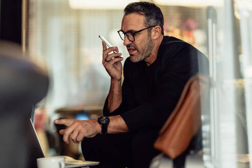 Reifer Geschäftsmann, der am Telefon spricht, während er auf seinen Laptop schaut. CEO arbeitet, während er im Hotelfoyer sitzt. - JLPSF18288