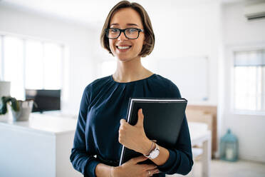 Lächelnde Geschäftsfrau mit Brille, die im Büro steht und ein Tagebuch hält. Porträt einer glücklichen Unternehmerin bei der Arbeit. - JLPSF18297