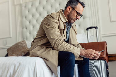 Porträt eines reifen Geschäftsmannes, der in einem Hotelzimmer sitzt. Ein Mann in formeller Kleidung sitzt auf dem Bett und benutzt ein Mobiltelefon. - JLPSF18304