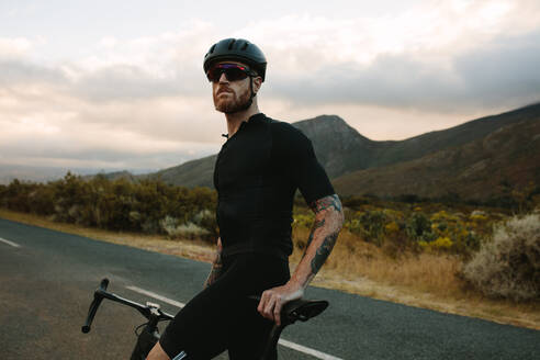 Porträt eines männlichen Radfahrers mit Helm und Sonnenbrille, der auf einer Landstraße steht und wegschaut. Männlicher Radfahrer, der auf einer leeren Landstraße eine Pause vom Radfahren macht. - JLPSF18309