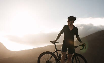 Pro bike rider taking break while riding in nature. Male cyclist standing with his bike on countryside road on sunny day. - JLPSF18330