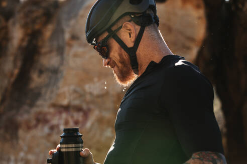 Radfahrer, der eine kurze Pause macht und Wasser trinkt, um während des Trainings hydriert zu bleiben. Sportler mit Fahrradhelm und Sonnenbrille, der beim Fahren im Freien Wasser aus einer Flasche trinkt. - JLPSF18340