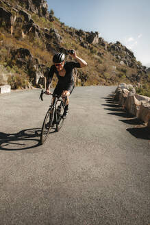 Sportler, der sich beim Radfahren auf einer Bergstraße Wasser über den Kopf gießt. Männlicher Radfahrer kühlt seinen Kopf beim Radfahren an einem Sommertag. - JLPSF18351